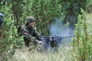 Vojinik u maskirnoj uniformi, sa nalepnicom zastave Srbije na desnom ramenu, ispaljuje granate koristeći Automatski bacač granata M93 na travnatoj površini